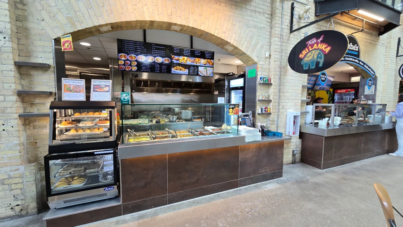 Taste of Sri Lanka counter inside The Forks Market, Winnipeg — hot curry service counter and pastry display under the arched heritage brickwork, with the hanging Taste of Sri Lanka elephant sign above